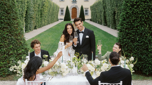 Elegant outdoor wedding reception with a bride and groom toasting with their guests at a beautifully decorated table surrounded by lush greenery and a stunning venue Villa Balbianoa at Lake Como in Italy in the background.