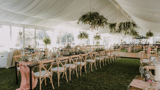 Elegant outdoor wedding reception setup in a draped tent with long wooden tables, greenery centerpieces, and hanging floral arrangements. Perfect inspiration for a romantic and natural wedding venue.