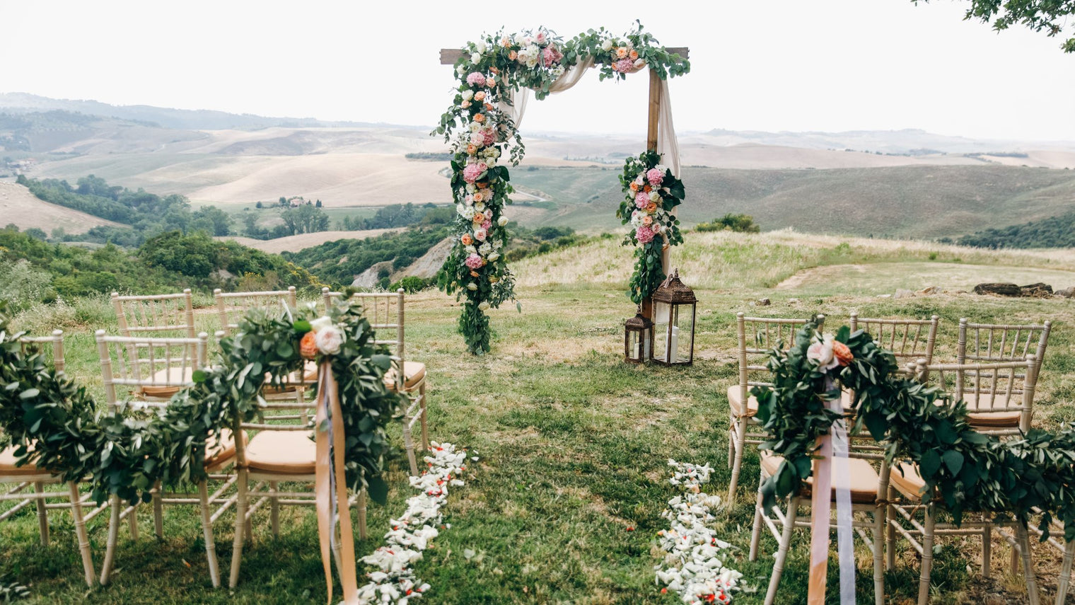 Outdoor wedding ceremony setup with floral-decorated arch, chairs with flower arrangements, and scenic countryside background.