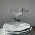 Load image into Gallery viewer, A customizable glass marker from the Champagne Collection, featuring a sleek black script-style name tag clipped to the rim of a crystal glass. The glass is elegantly placed on a stack of white plates, adorned with a black ribbon tied around its stem, showcasing the sophisticated and personalized design of the place tag.
