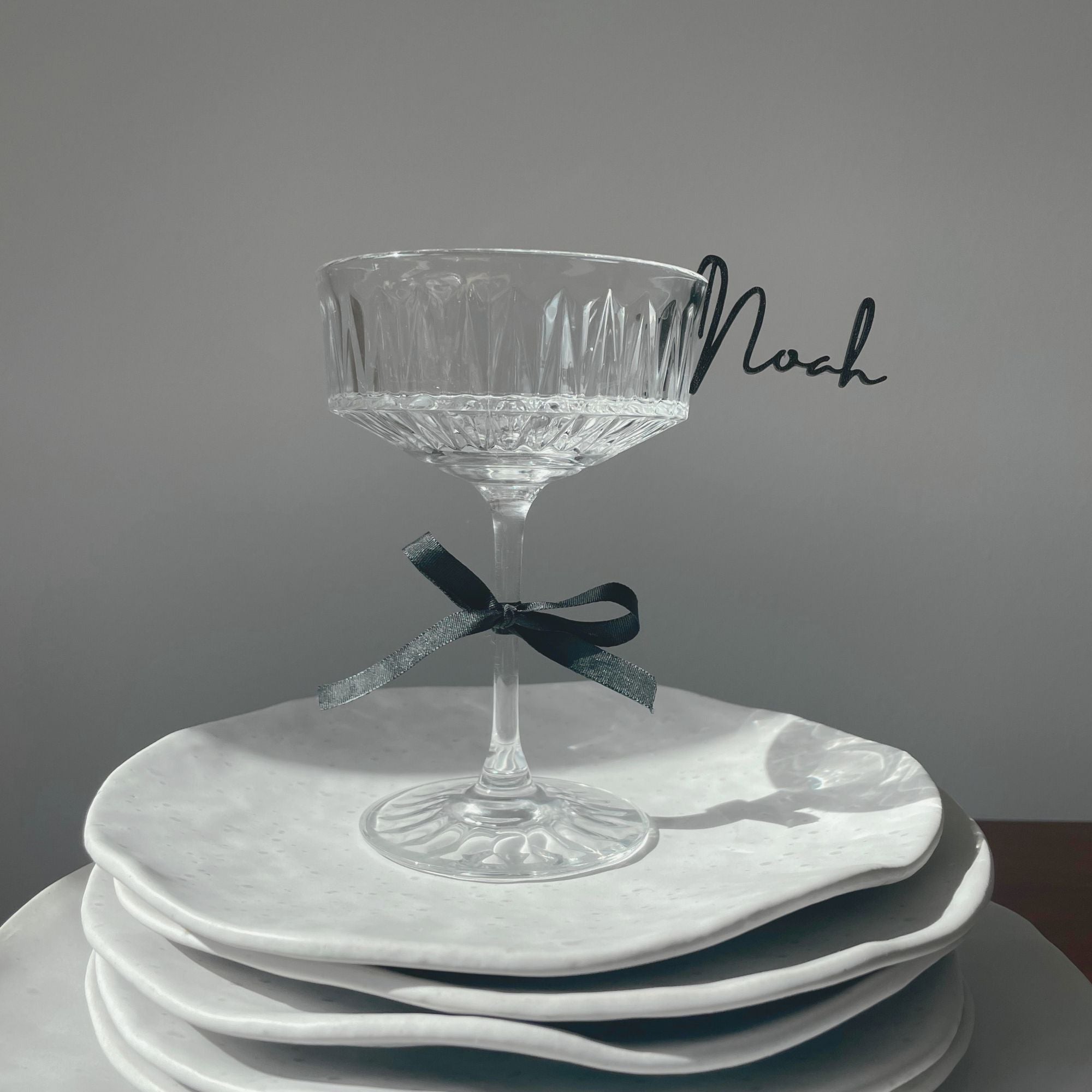 A customizable glass marker from the Champagne Collection, featuring a sleek black script-style name tag clipped to the rim of a crystal glass. The glass is elegantly placed on a stack of white plates, adorned with a black ribbon tied around its stem, showcasing the sophisticated and personalized design of the place tag.