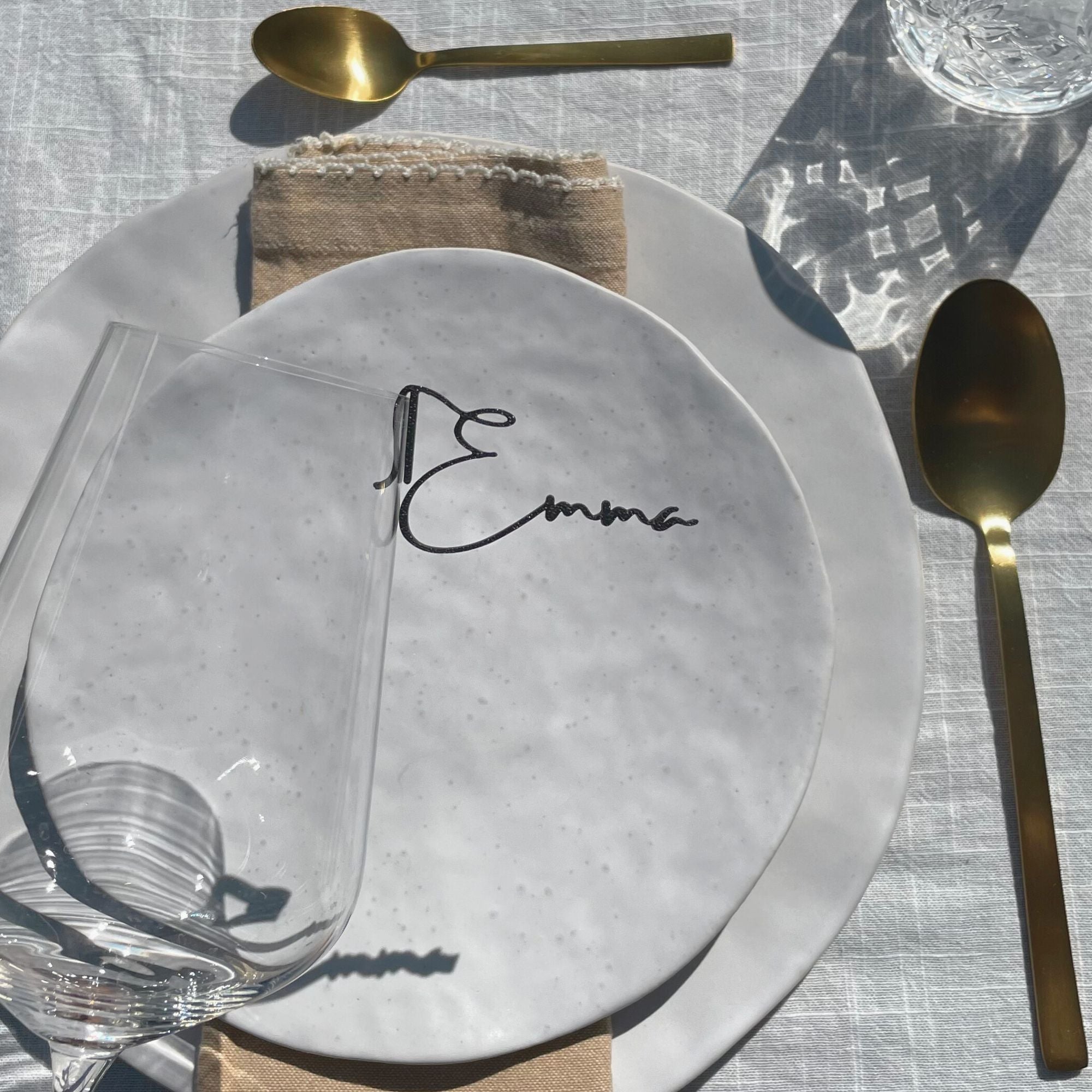 A beautifully set table featuring a white plate with a customizable name tag saying "Emma" from the Champagne Collection from Cheers Day. The setup includes a gold spoon, a beige napkin, and a wine glass with a "Cheers" drink topper. The glass charm elegantly marks the wine glass, showcasing the sophisticated and personalized touch of the place card in the tablescape.