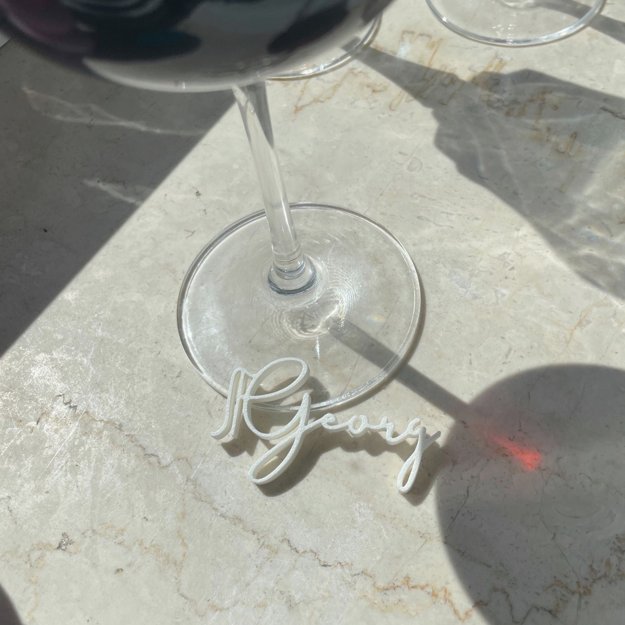 Close-up view of a customizable wine glass marker with the name "Georg" from the Champagne Collection by Cheers Day. The elegant place card alternative is shown attached to the base of a wine glass, highlighting its sophisticated and functional design.